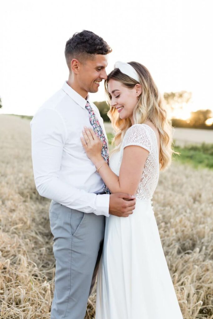 Bride and groom stand together in a field as the sun goes down in the background.